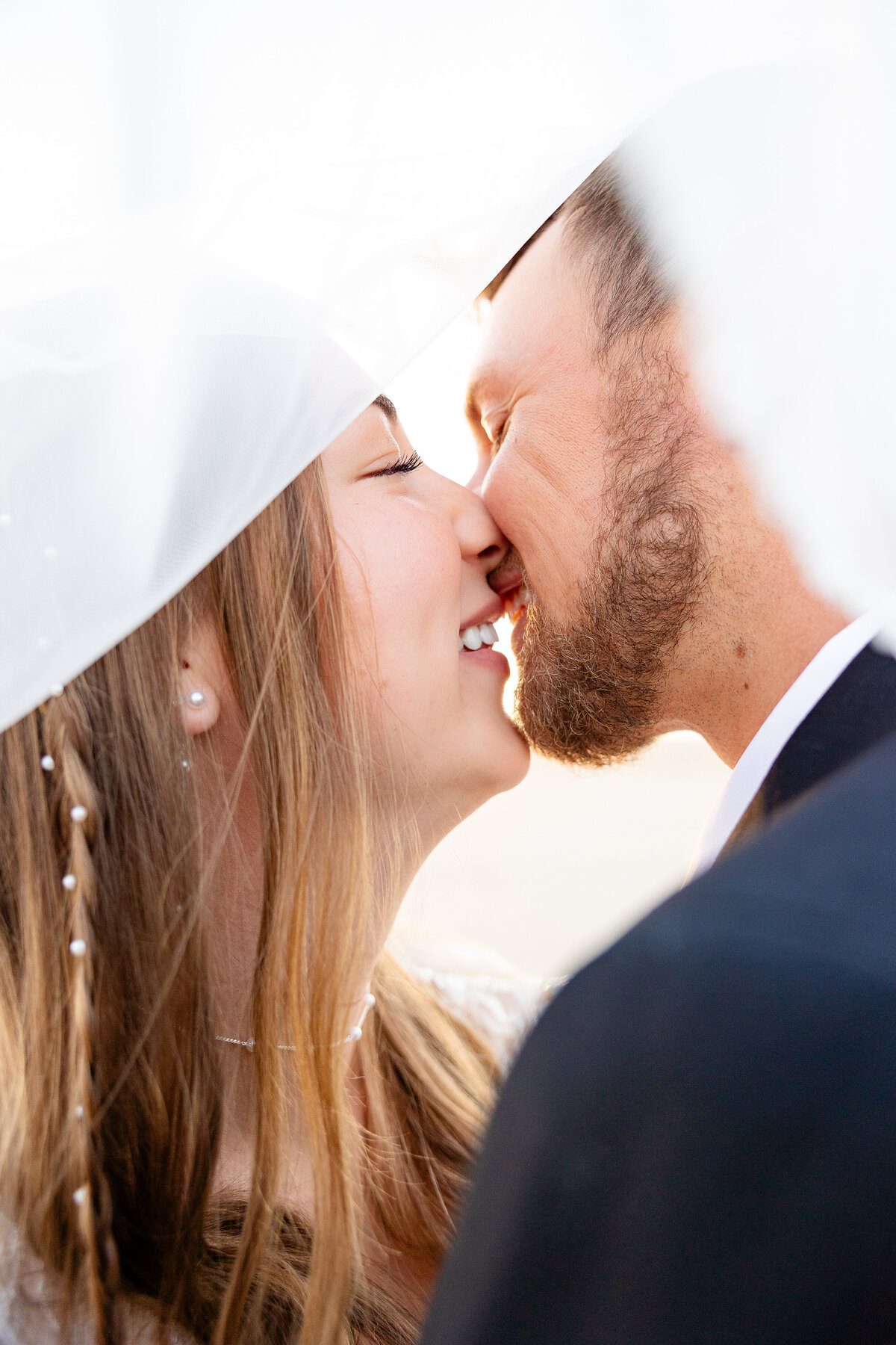 Close-up of a bride and groom kissing under the bride's veil, illuminated by soft morning light.. Captured by Cassie Lee Photography