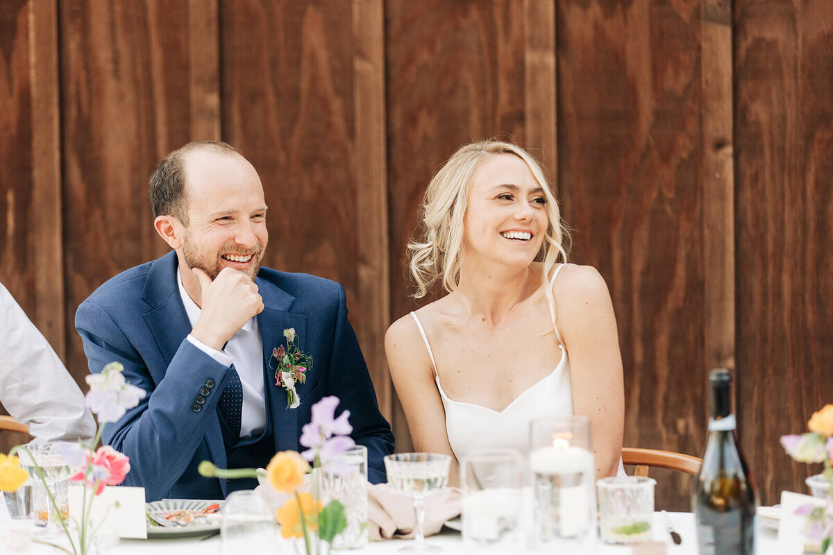 A bride and groom seated at an outdoor table smiling.