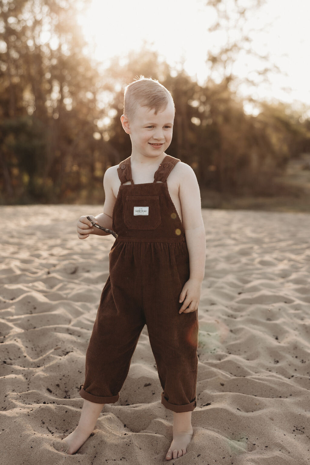 Young child wearing brown overalls stands barefoot on sandy ground outdoors, holding an object in one hand with trees visible in the background—a perfect moment captured by a NSW family photographer.