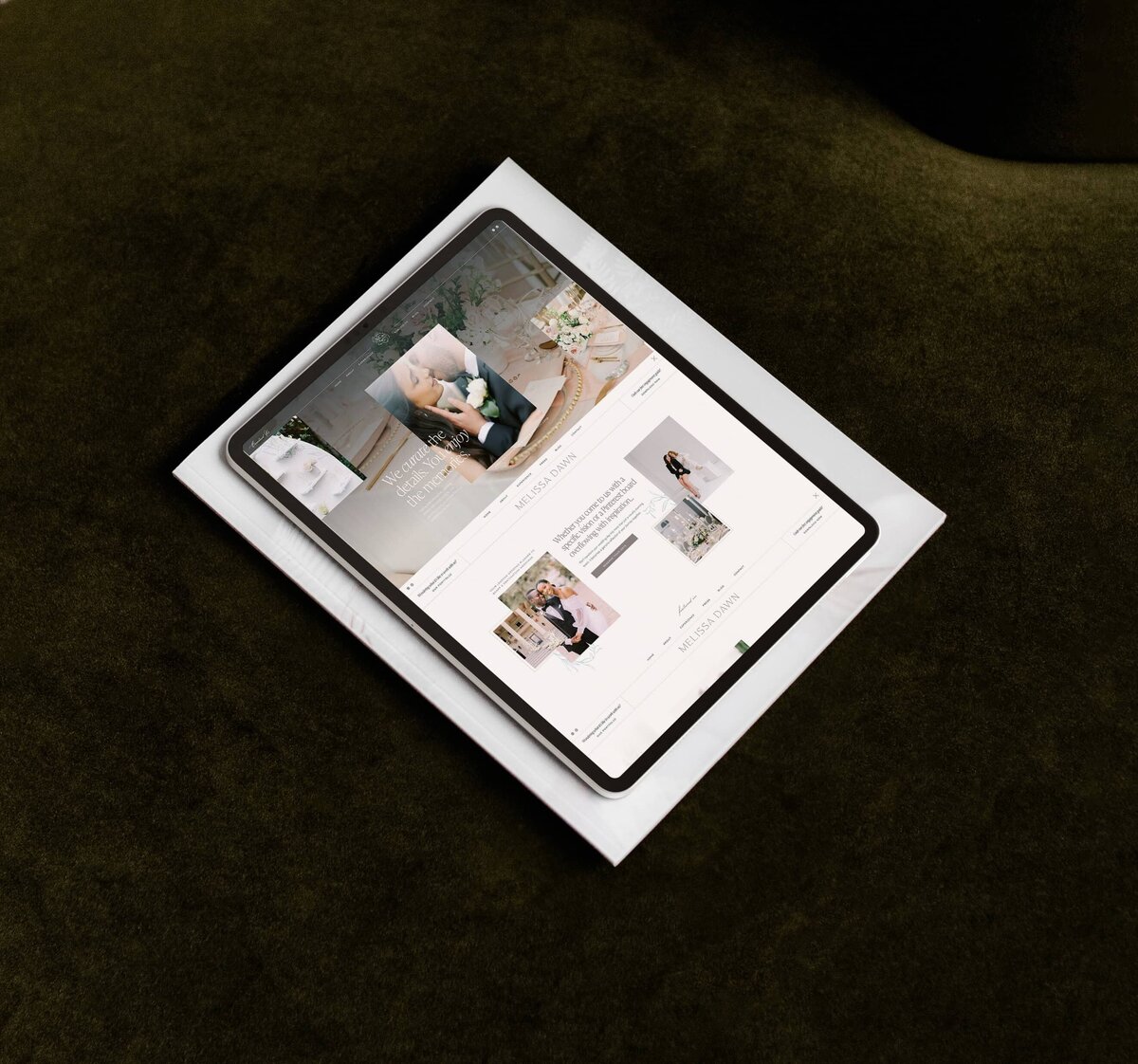 A tablet displaying a wedding planner logo and website sits on top of a closed hardcover book, placed on a dark green surface.