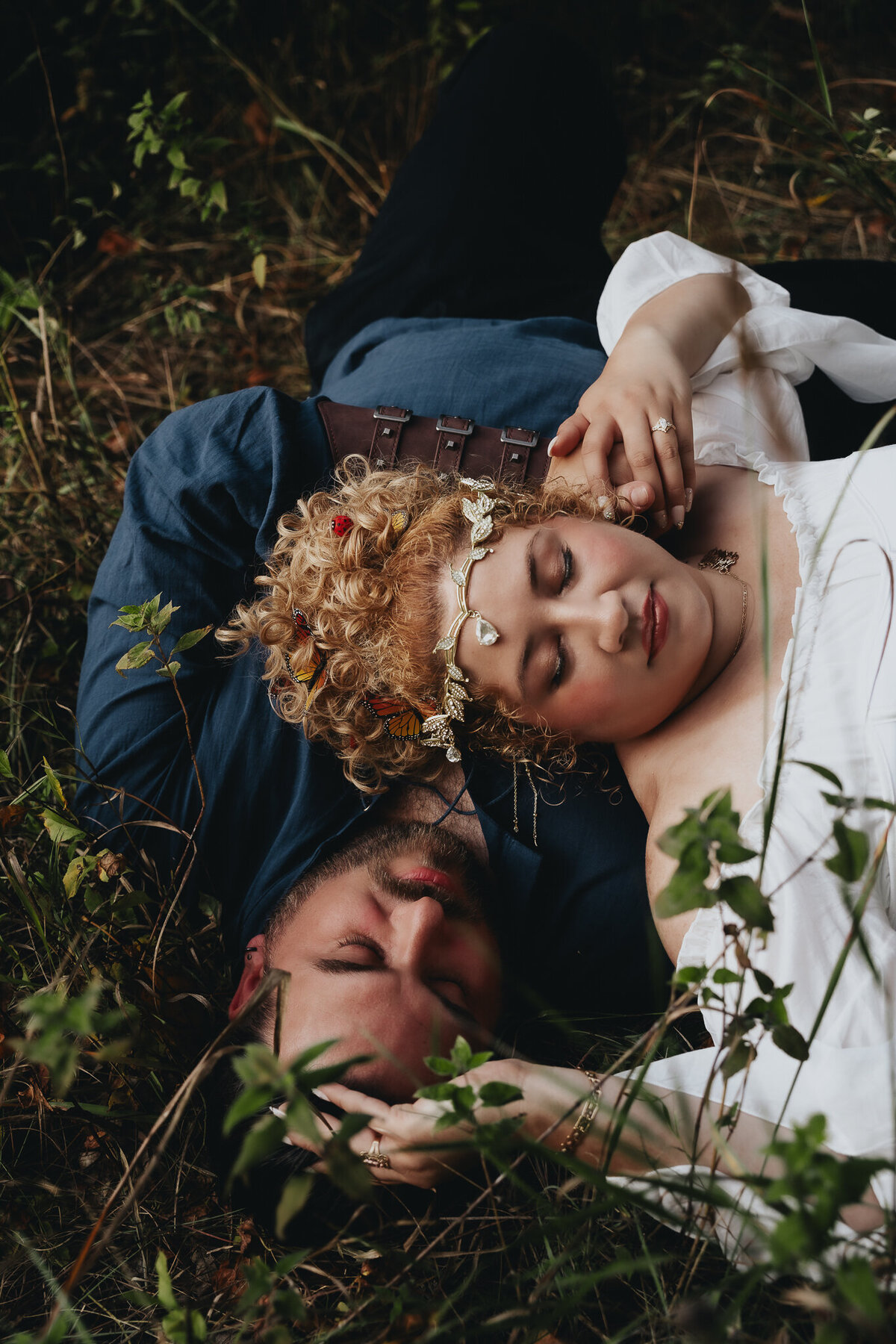 Couple dressed in fantasy-inspired outfits lies in the grass with crowns and accessories during creative couples photoshoot.
