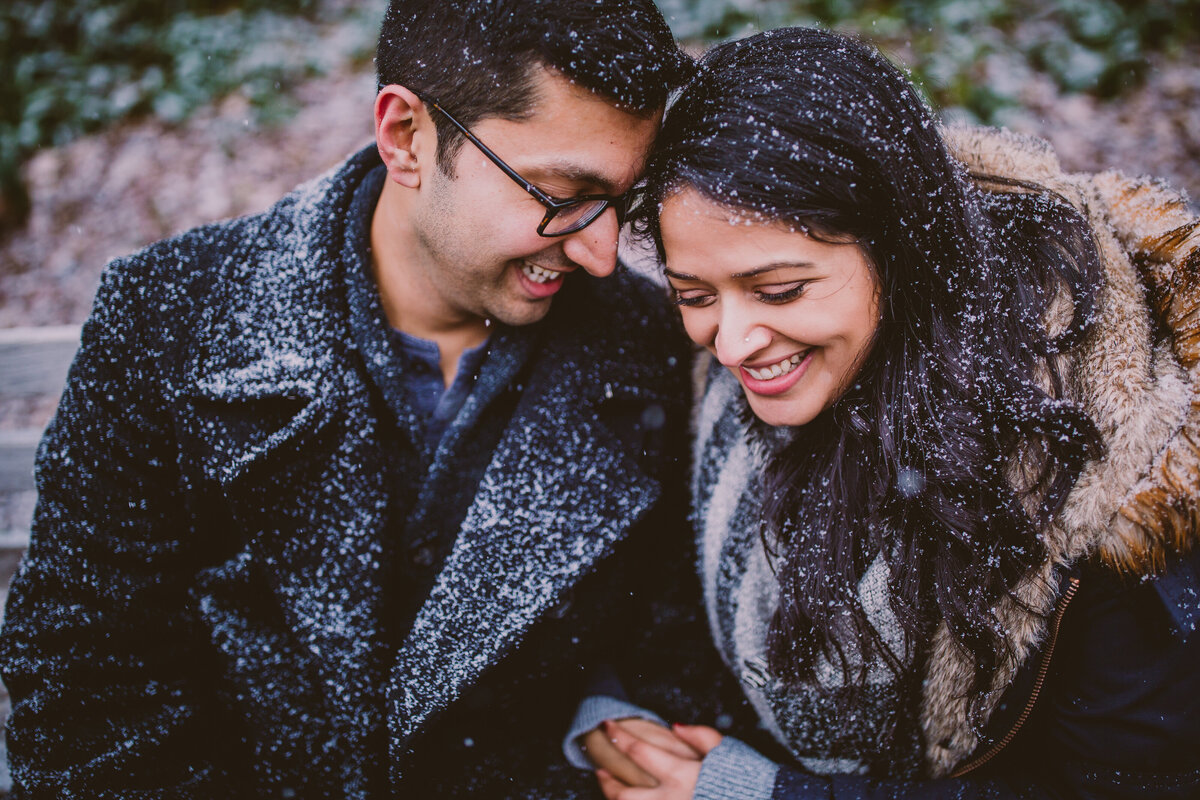 A couple during their winter engagement session sitting and sharing a moment at a park during a snowstorm with snowflakes all over them 