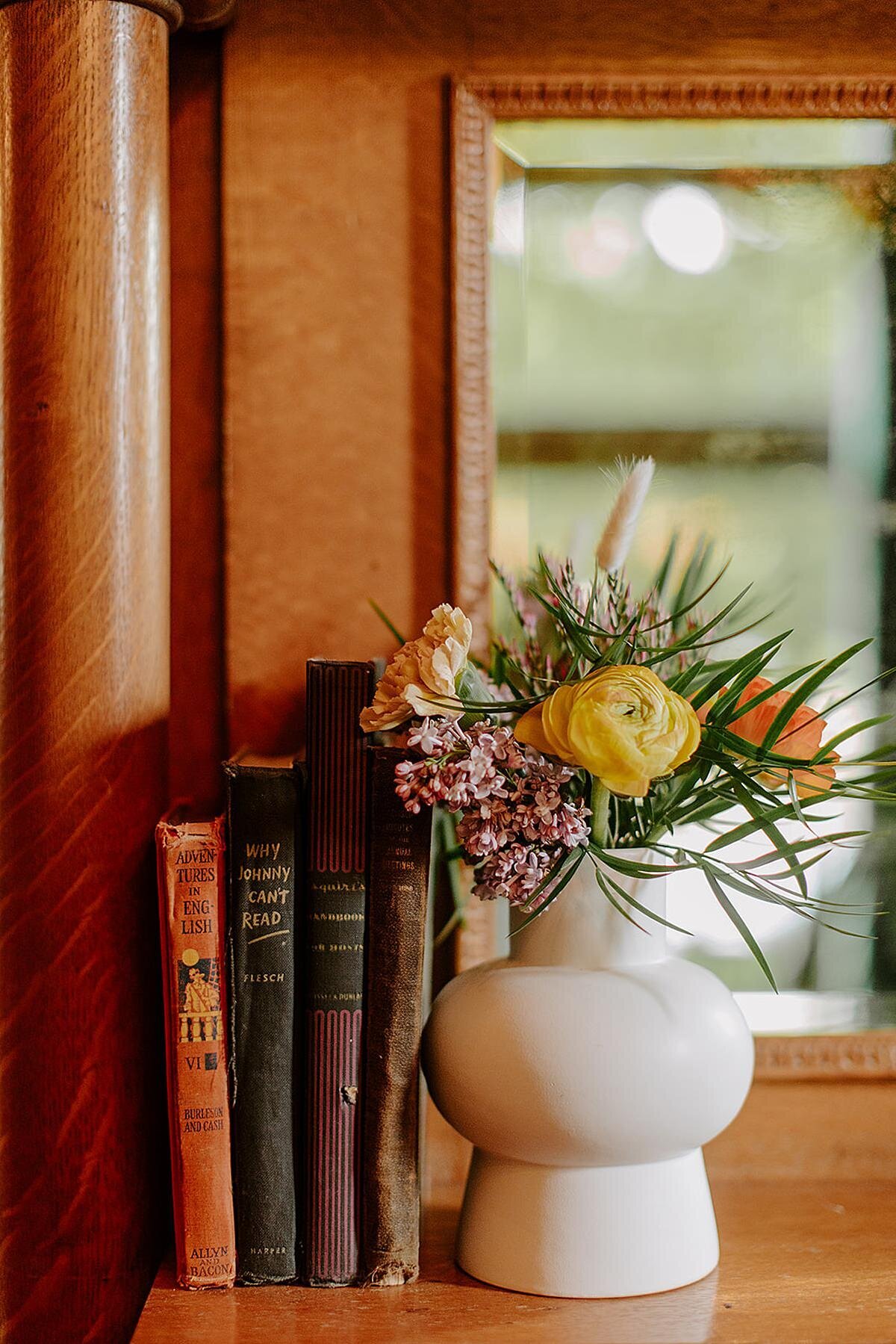 A rounded white ceramic vase sits on a light wood window sill propping up four vintage books. The vase has purple wax flower, blush carnations, yellow ranunculus, and orange ranunculus with willow and greenery.