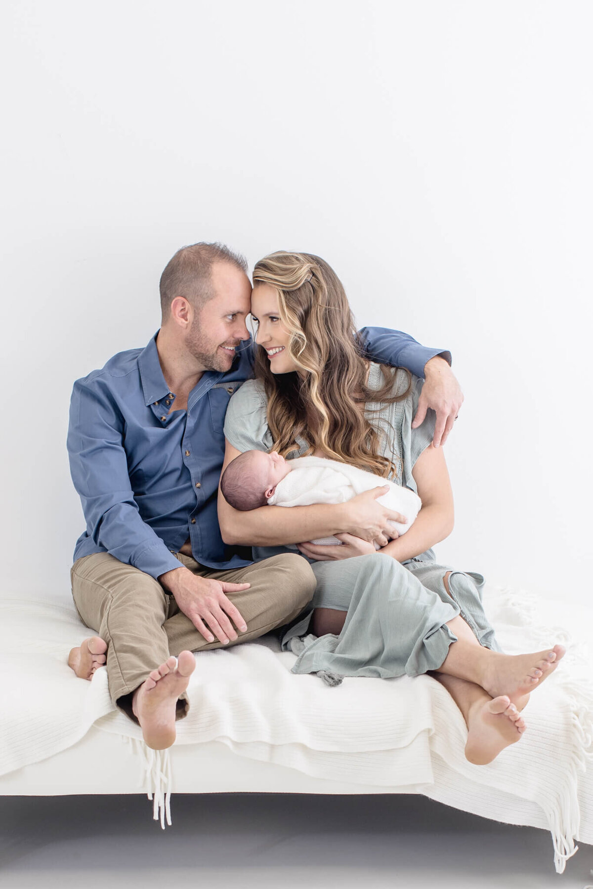 A couple sits closely on a white bed, smiling at each other. The woman cradles a swaddled newborn in her arms. Both are barefoot, with the man wearing a blue shirt and khaki pants, and the woman in a light green dress.