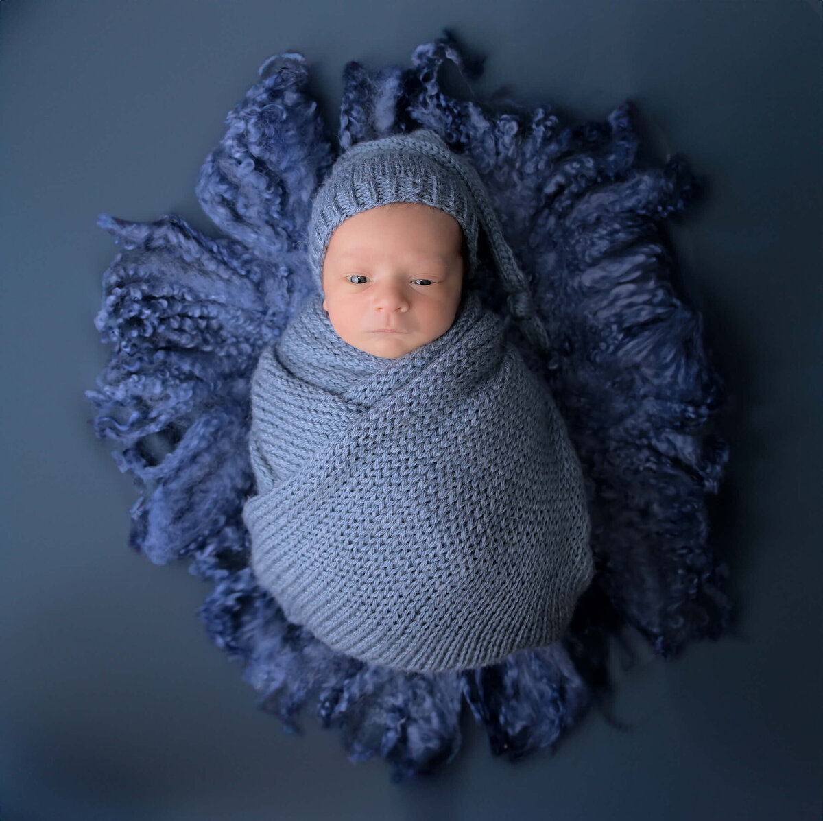 Beth Benecki of E. Benecki Photography captures a newborn baby boy wrapped in blue knit wrap and placed on similar shades of textured fabrics. Baby is photographed  with his eyes wide open