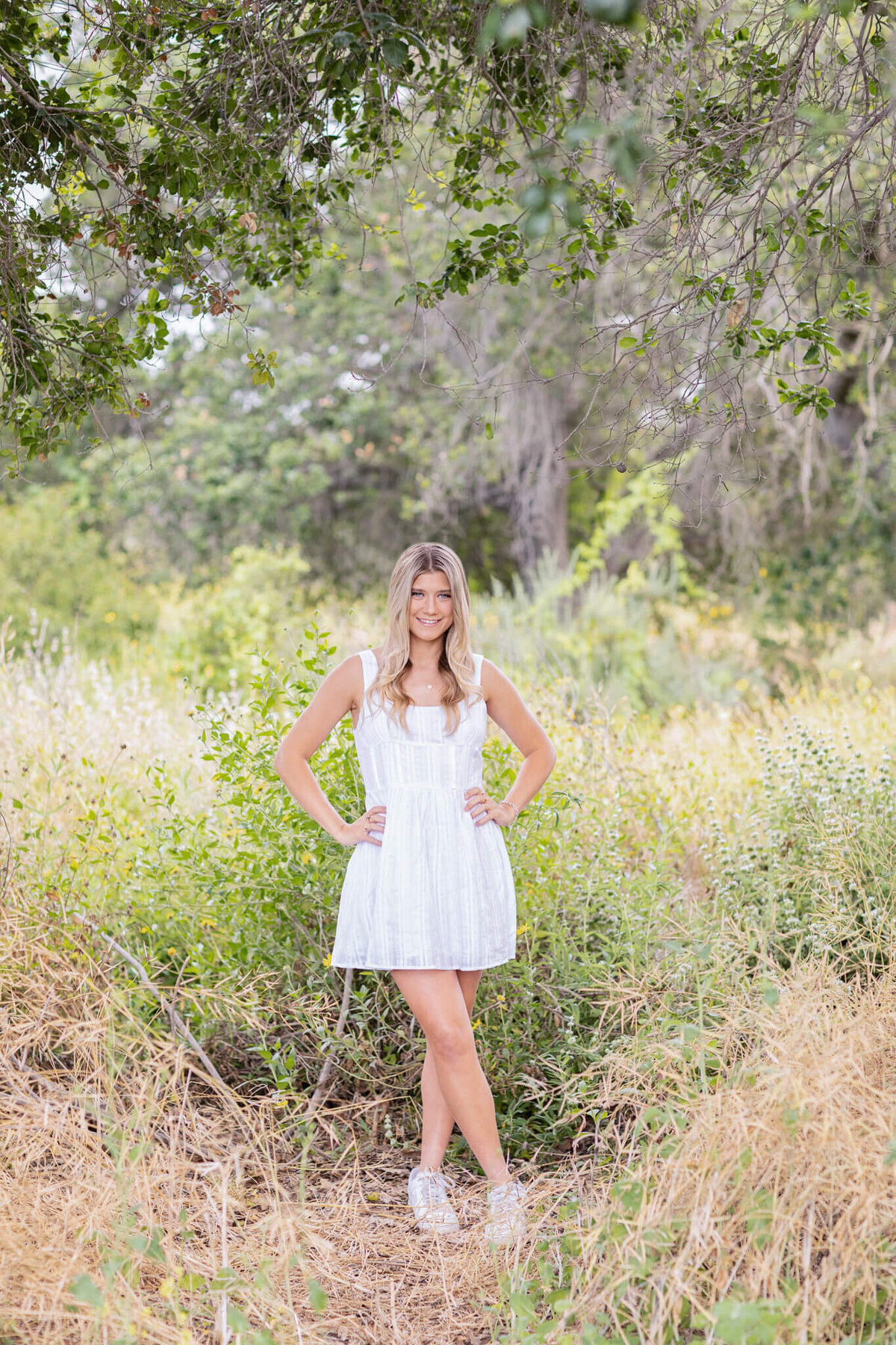 A woman stands in a grassy, wooded area wearing a white dress and sneakers. She is smiling with her hands on her hips. The background features green foliage and trees.