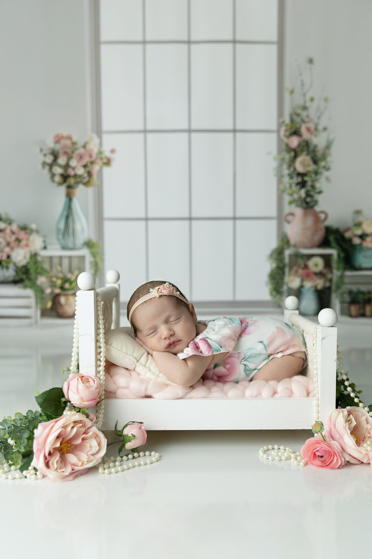 A newborn baby sleeps in a floral print onesie on a tiny wooden bed with pearls and flowers under a window in a studio of a NJ Newborn Photography