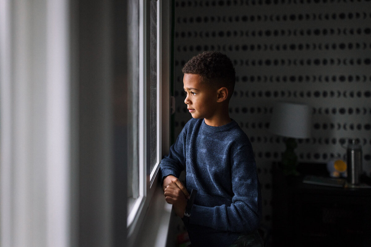 8-year-old boy standing by window looking out with a serious expression