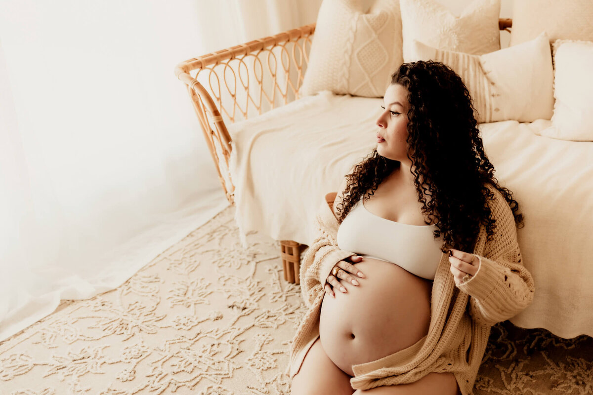 Woman in her third trimester looking out of a window while playing with her hair and holding her baby bump. 
