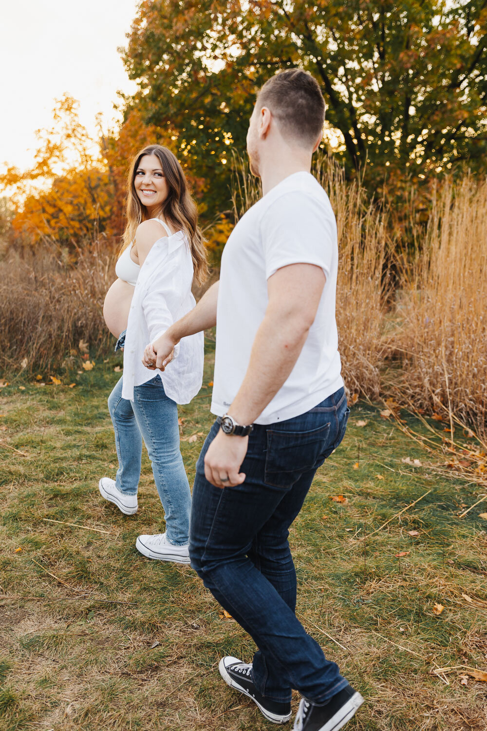 Expecting woman casually dressed in jeans and an open white button up shirt, leads her husband.  She is holding his hand and looking back at him.