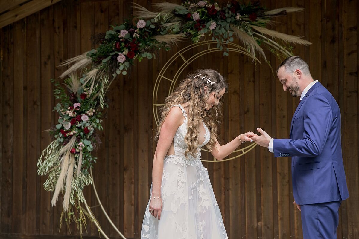 The bride, wearing a long, sleeveless lace wedding gown with a plunging neckline holds hands with the groom, wearing a bright blue suit for their first look underneath their boho ceremony arbor at the rustic wooden pavilion at Saddle Woods Farm