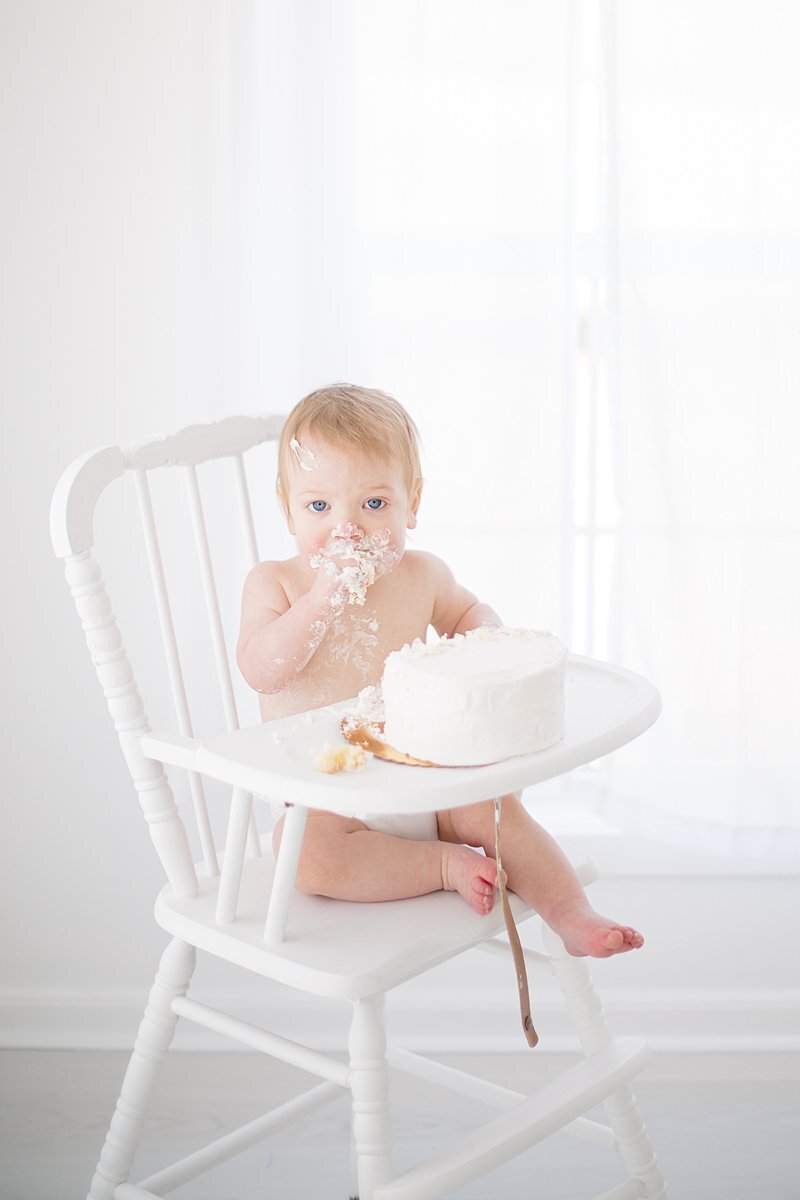 Baby eating cake sitting in white high chair for 1st birthday photos