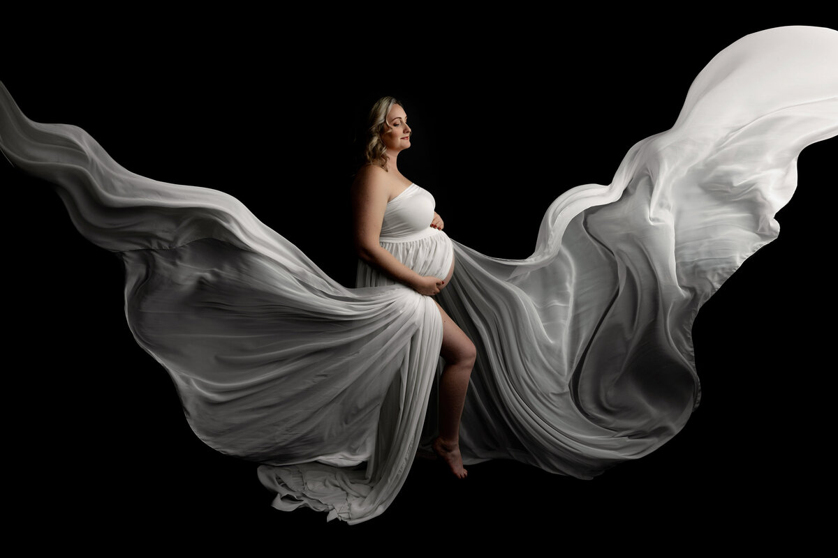 A mother to be in a white silk maternity gown smiles as her long train flows into the air around her