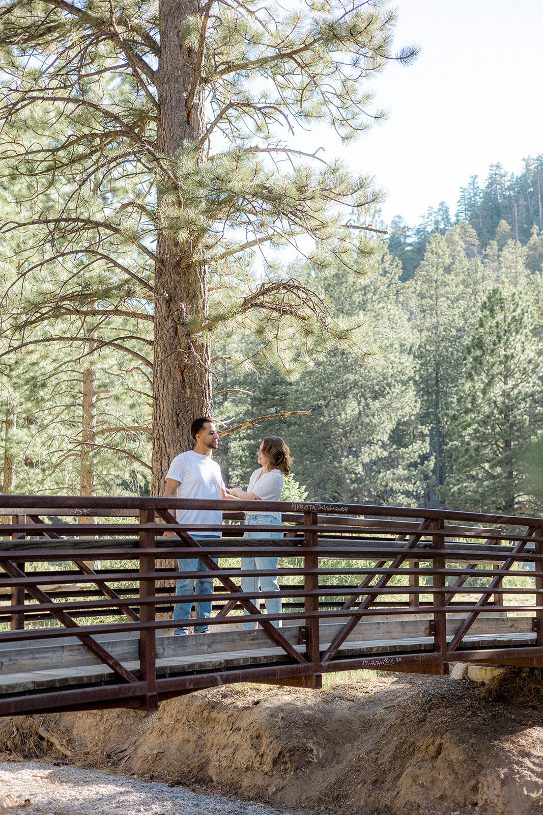 A couple stands on a wooden bridge in the forest, looking at each other with soft smiles, surrounded by tall pine trees.