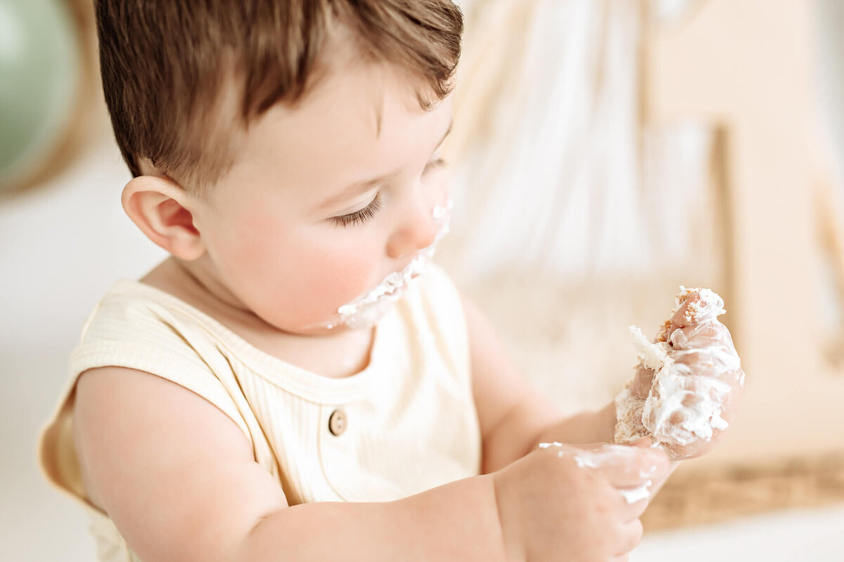 A baby with cake frosting smeared on their face and hands is intently examining a small piece of cake. The baby is wearing a light-colored outfit and is surrounded by a softly blurred background.