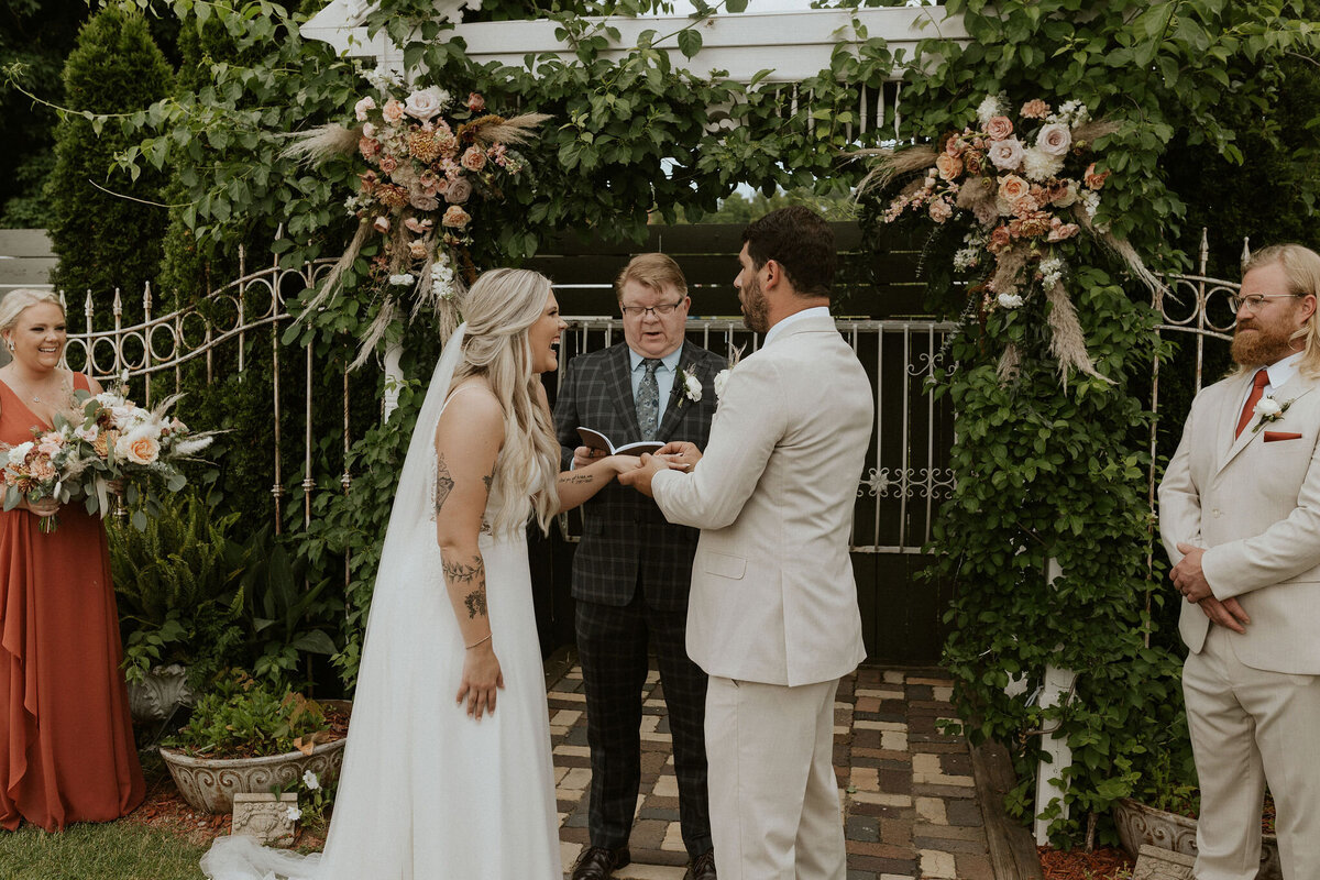 Bride and groom standing at an ivy covered alter laughing as groom puts wedding band on bride's finger