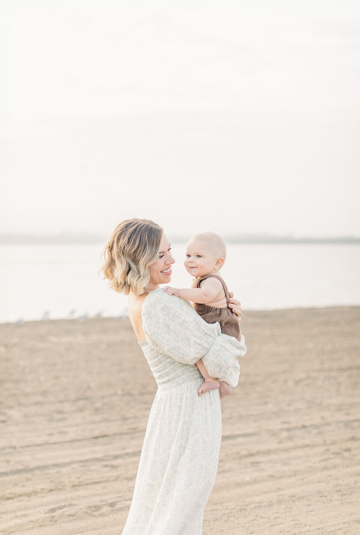 Mother in a flowing dress stands barefoot on the sandy shore holding her baby boy during a 6-month milestone session at a Kansas City lake.
