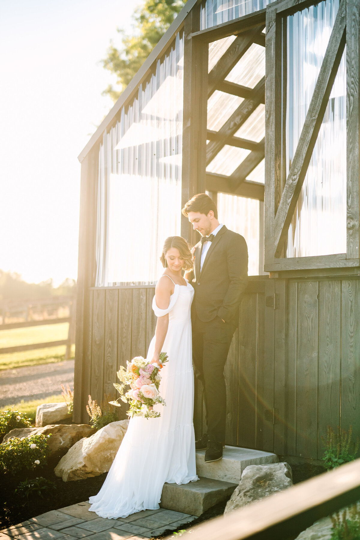 a bride in a white gown with pink and white flowers and a groom in a black suit in front of the garden house at Willowbrook wedding venue in the sunset