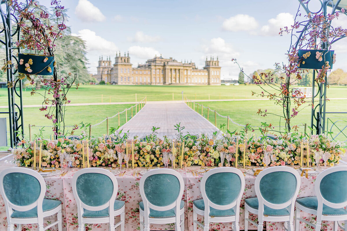 a long wedding dinner table with pink and peach flowers and blue chairs overlooking blenheim palace for a luxury wedding by westacott weddings