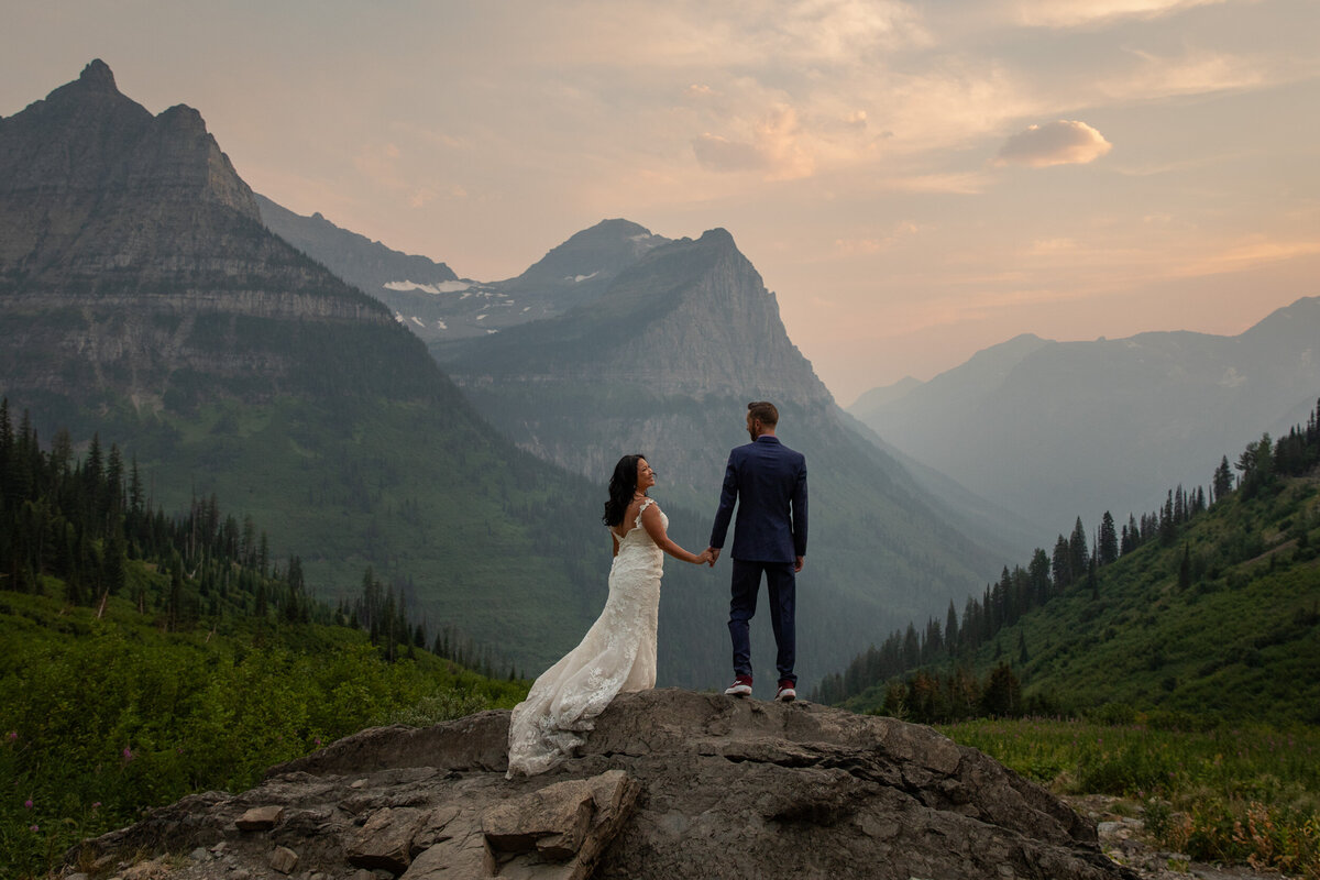 Bride in a lace wedding dress and groom in a navy blue suit stand on a rocky mountain ridge, holding hands and looking out over a dramatic 