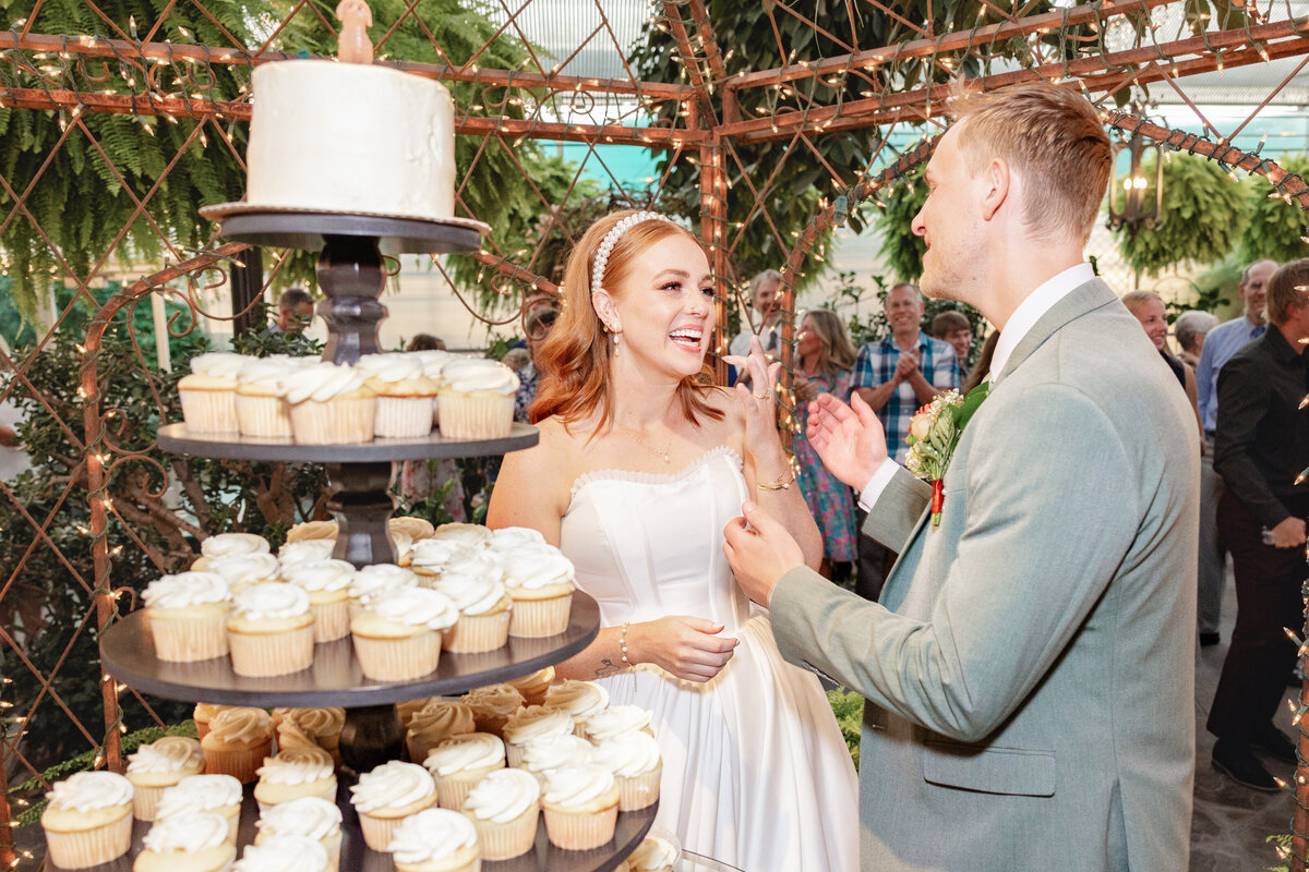 The bride and groom smile and laugh as they prepare to cut their wedding cake, surrounded by an elegant cupcake display and glowing fairy lights.