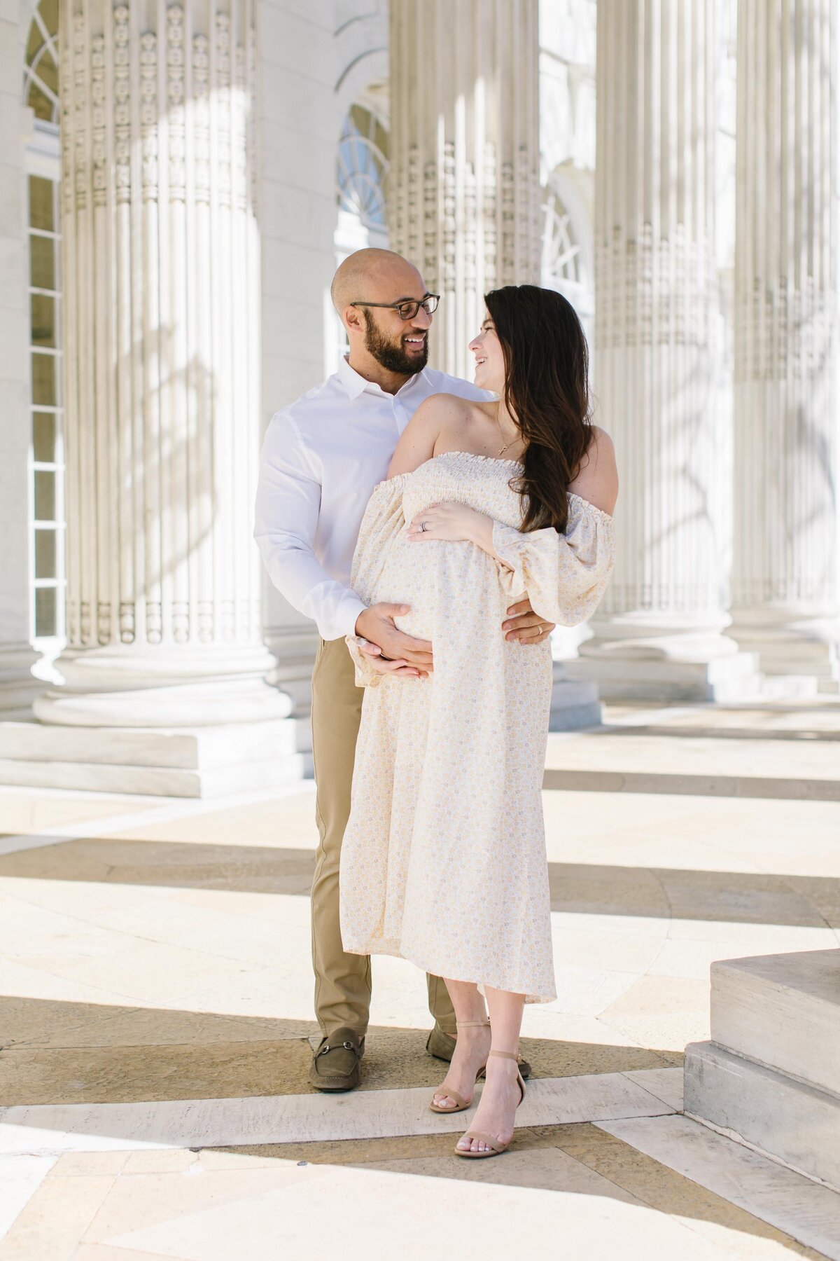 Woman holding baby bump with husband smiling behind her during photography session