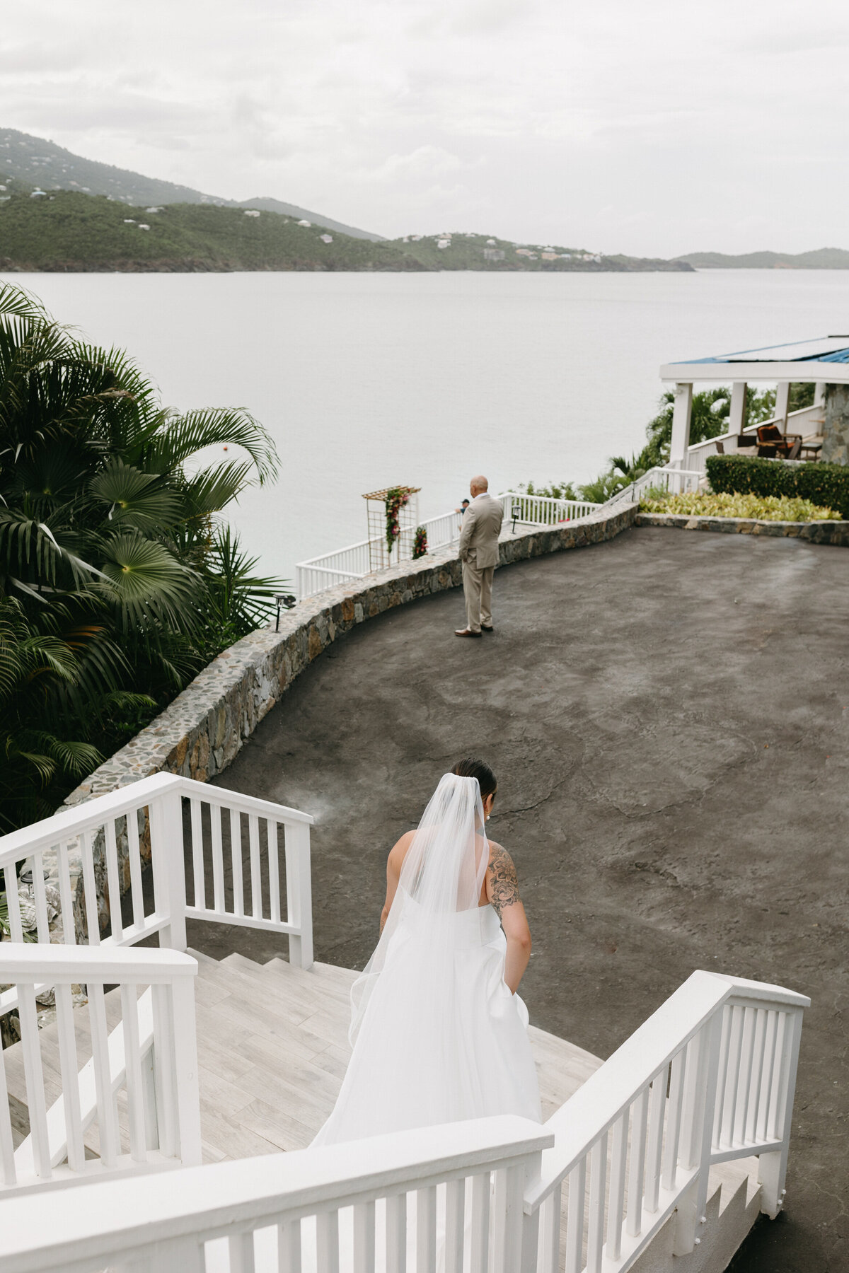 A bride descending outdoor stairs towards a waiting groom against a backdrop of a tropical seaside resort
