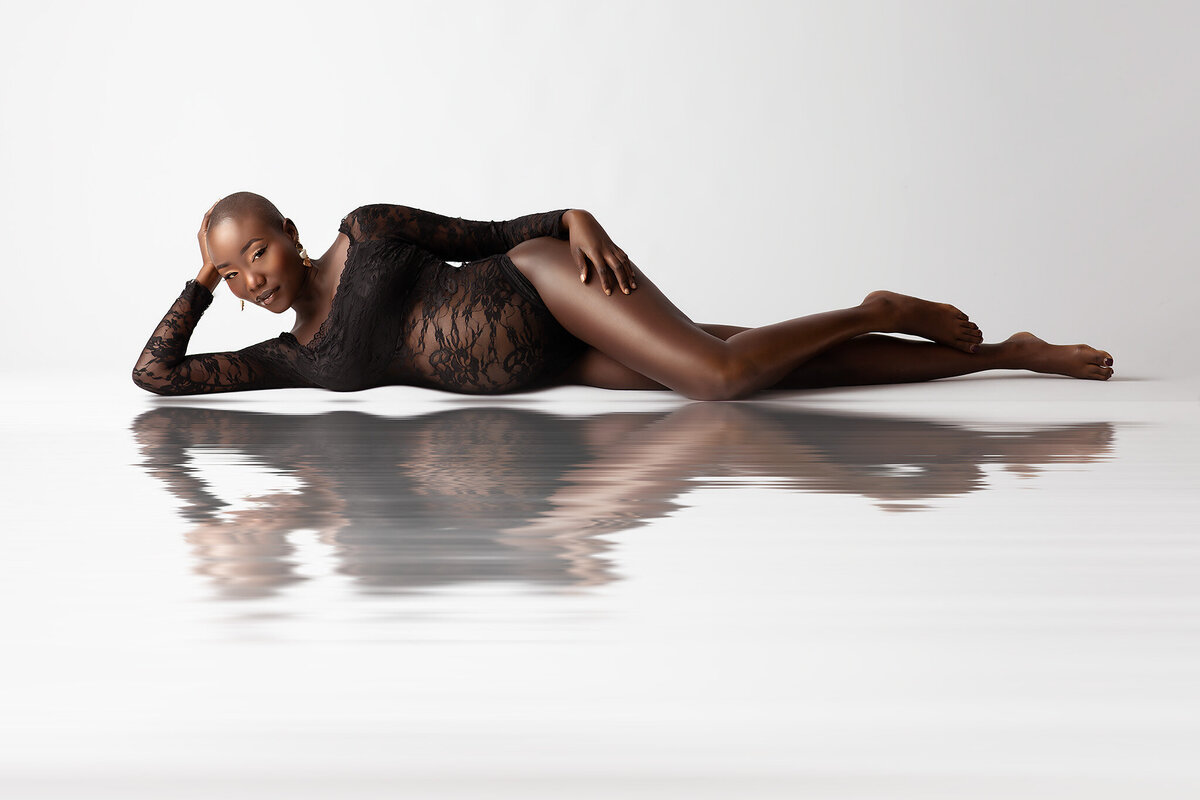 A serene maternity photograph of a woman reclining gracefully on a reflective surface, dressed in a sheer black lace bodysuit. The minimalist white background and elegant pose highlight her natural beauty, skillfully captured by Lindsay Walden, the best of Dallas maternity photographers.