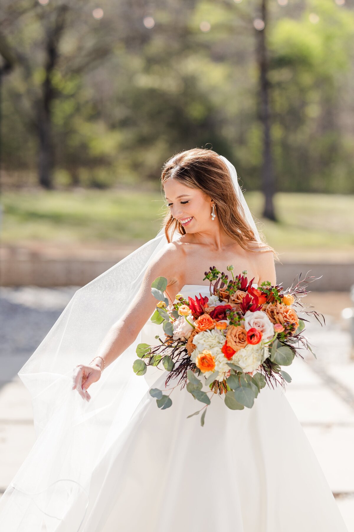 An image captures by an Arkansas wedding photographer of a bride playing with her veil in the sun while holding a large colorful bouquet