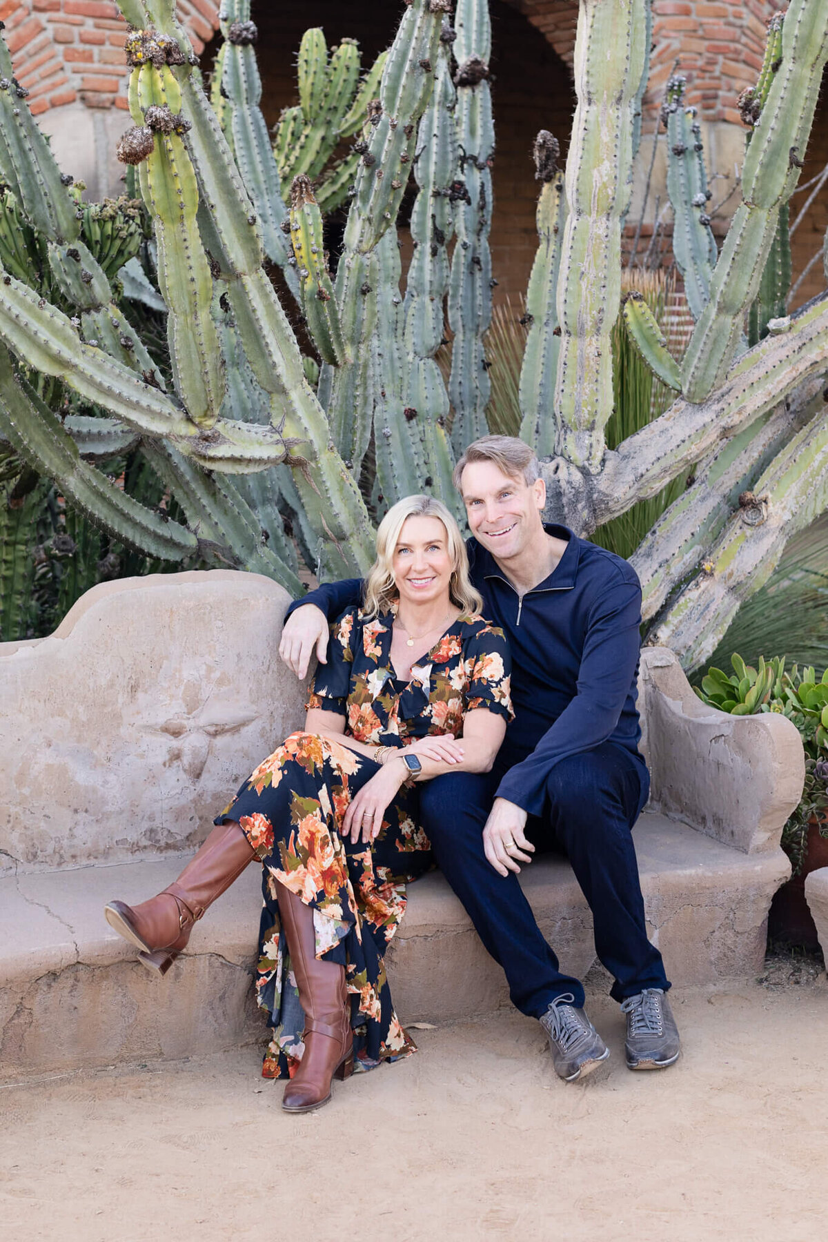 A woman with blonde hair wearing an orange and navy dress and high brown boots, leaning against her husband's chest, wearing a navy long sleeve shirt, as they're sitting on a clay bench with large cactus plants behind them.