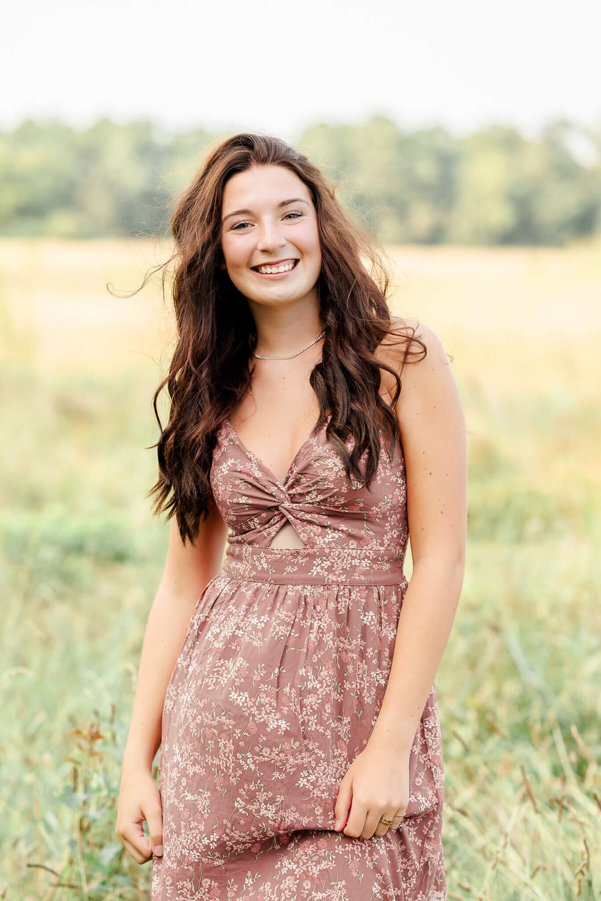A high school senior, wearing a long floral dress, stands in the tall grass at Bells Mill Park in Chesapeake.