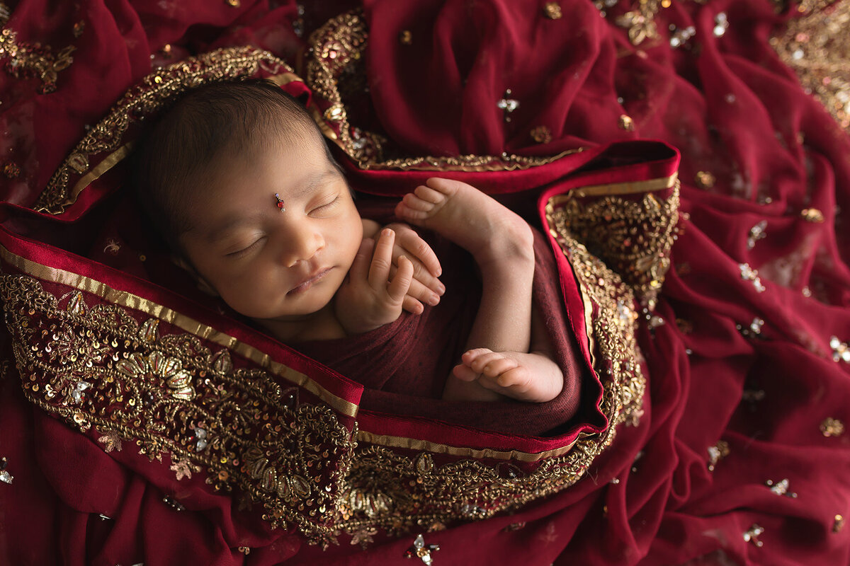 Newborn baby girl wrapped in a richly embroidered red sari with gold details, featuring a bindi on her forehead, photographed by Dallas newborn photographer.