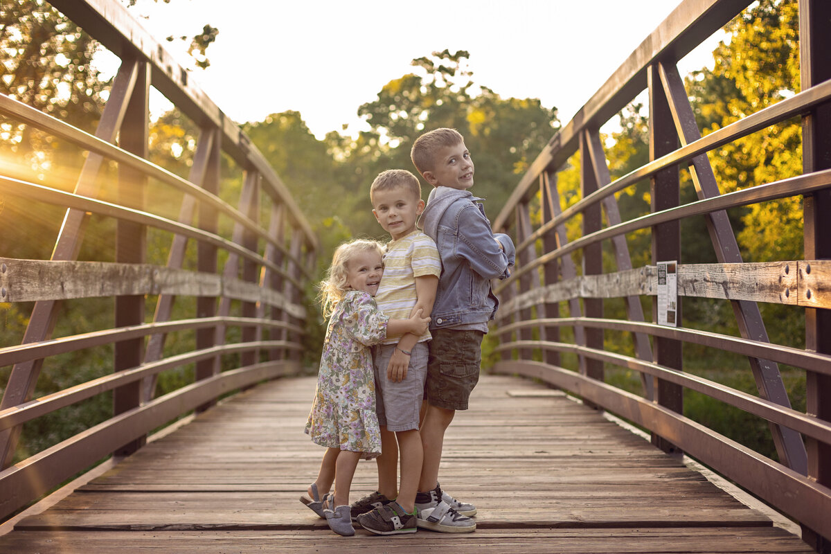 Two toddler brothers stand back to back while their younger sister hugs one of them as they stand on a bridge at sunset for a New Jersey Family Photographer