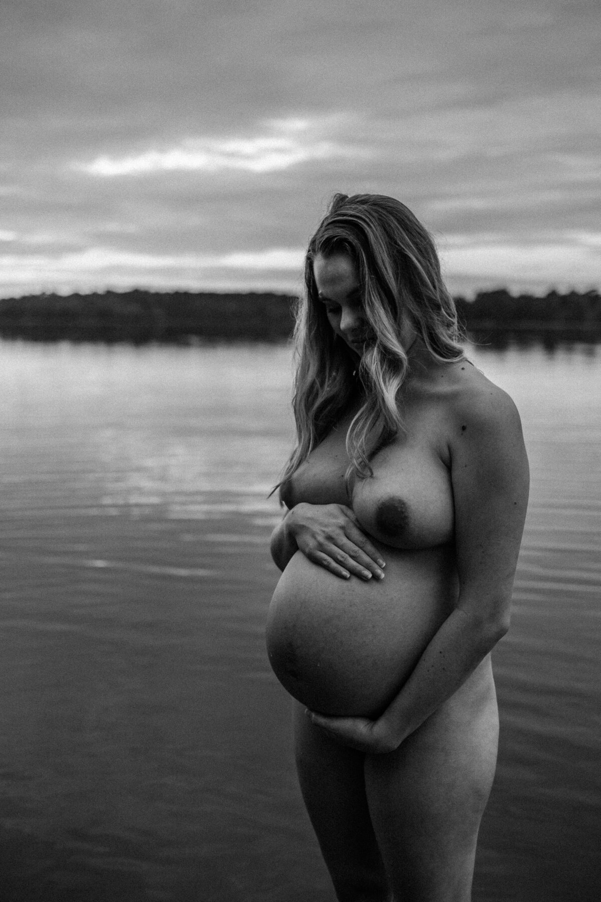A pregnant woman stands unclothed near a calm body of water, with one hand on her belly and another on her chest. The scene is in black and white and captures the serene outdoor environment. Durham Boudoir Photography