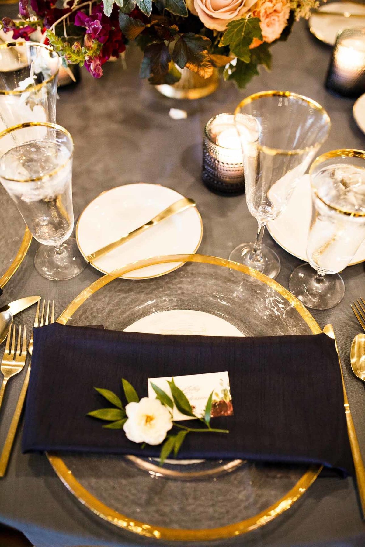 wedding reception place setting with navy blue linen, wrapped around gold rimmed glass charger lpate, with single flower on napkin