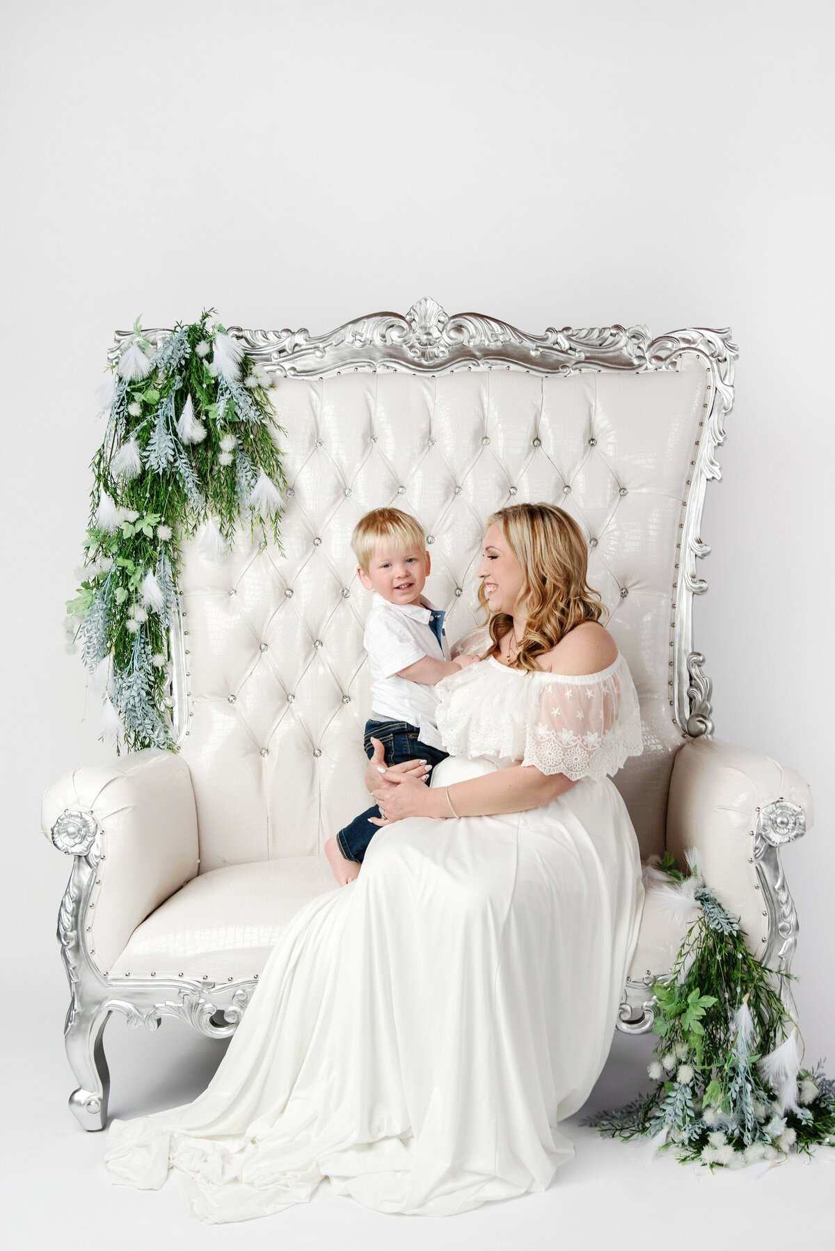 st-louis-family-photographer-mom-in-white-dress-looking-at-toddler-son-sitting-on-white-throne-with-flowers