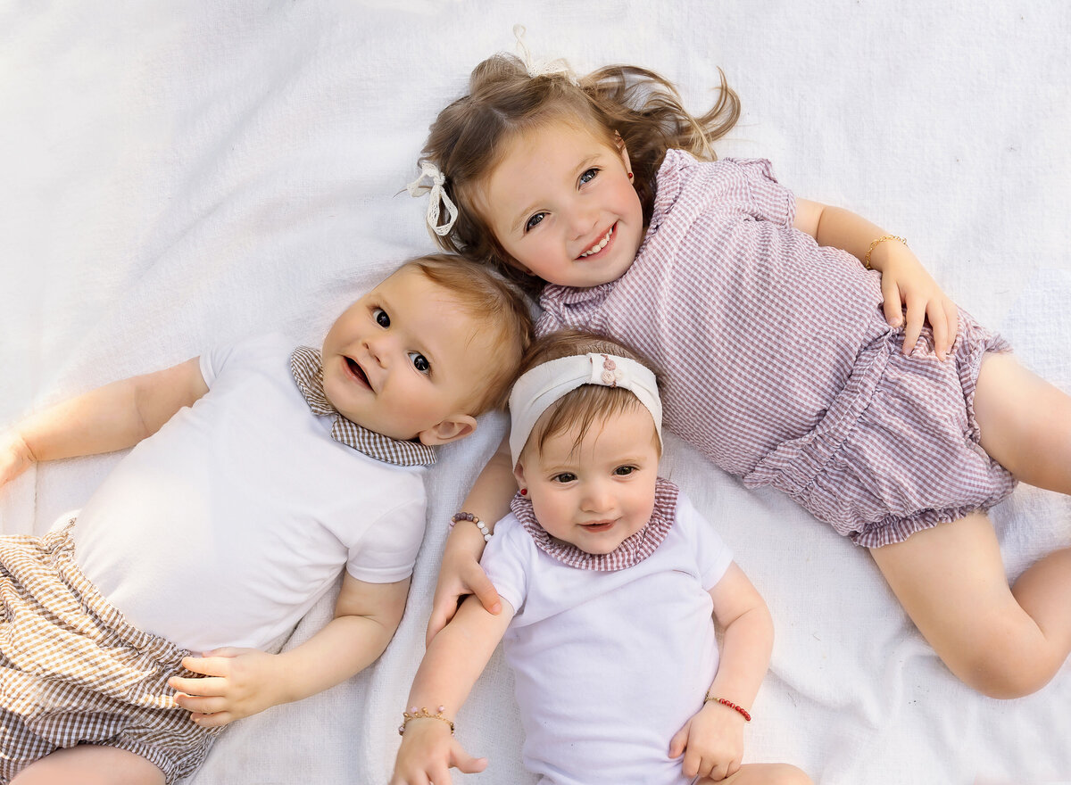 Three siblings are laying on a blanket for a family photoshoot in brooklyn, ny. All are looking up at the camera and smiling. Captured by brooklyn family photographer Chaya Bornstein
