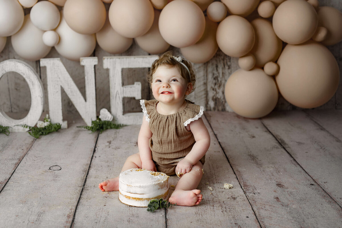 one year old girl wearing a brown outfit with ruffles sitting in front of a white birthday cake with white one letters and neutral colored balloons at a cake smash session