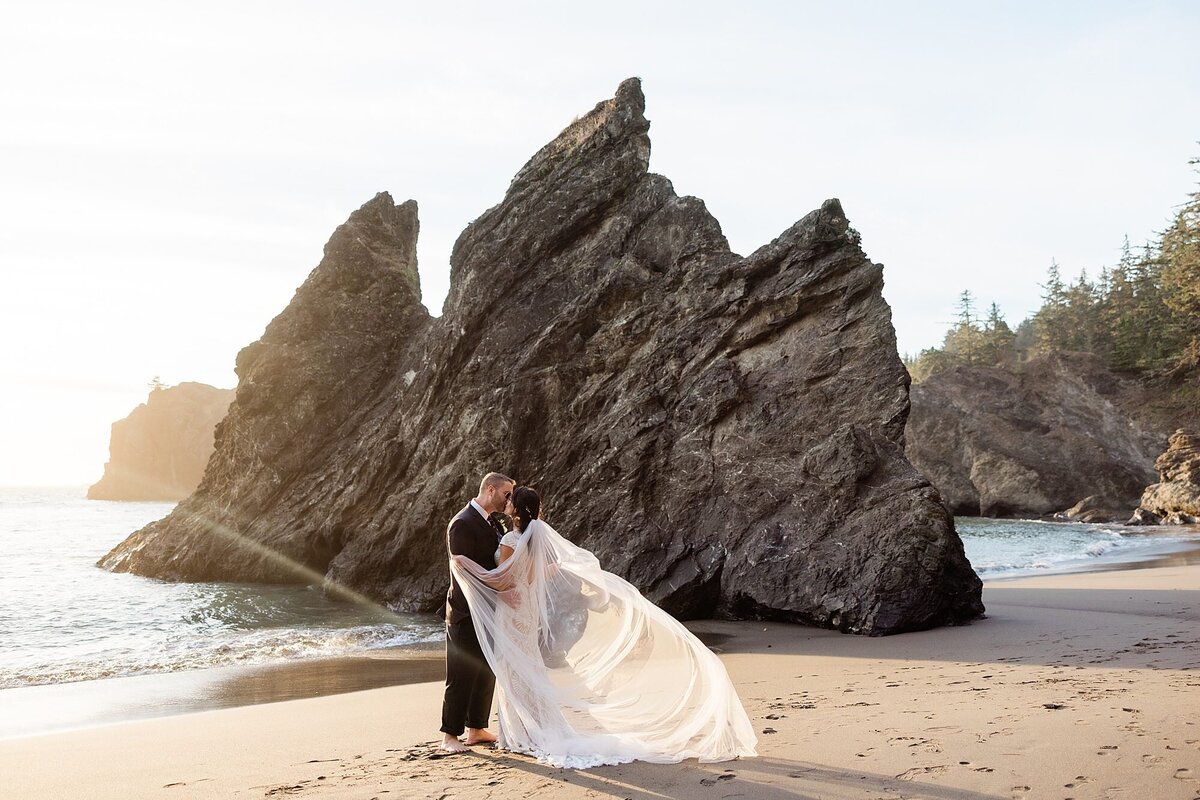A bride with her long veil stands on the beach and kisses the groom in front of large sea stacks and rock formations.