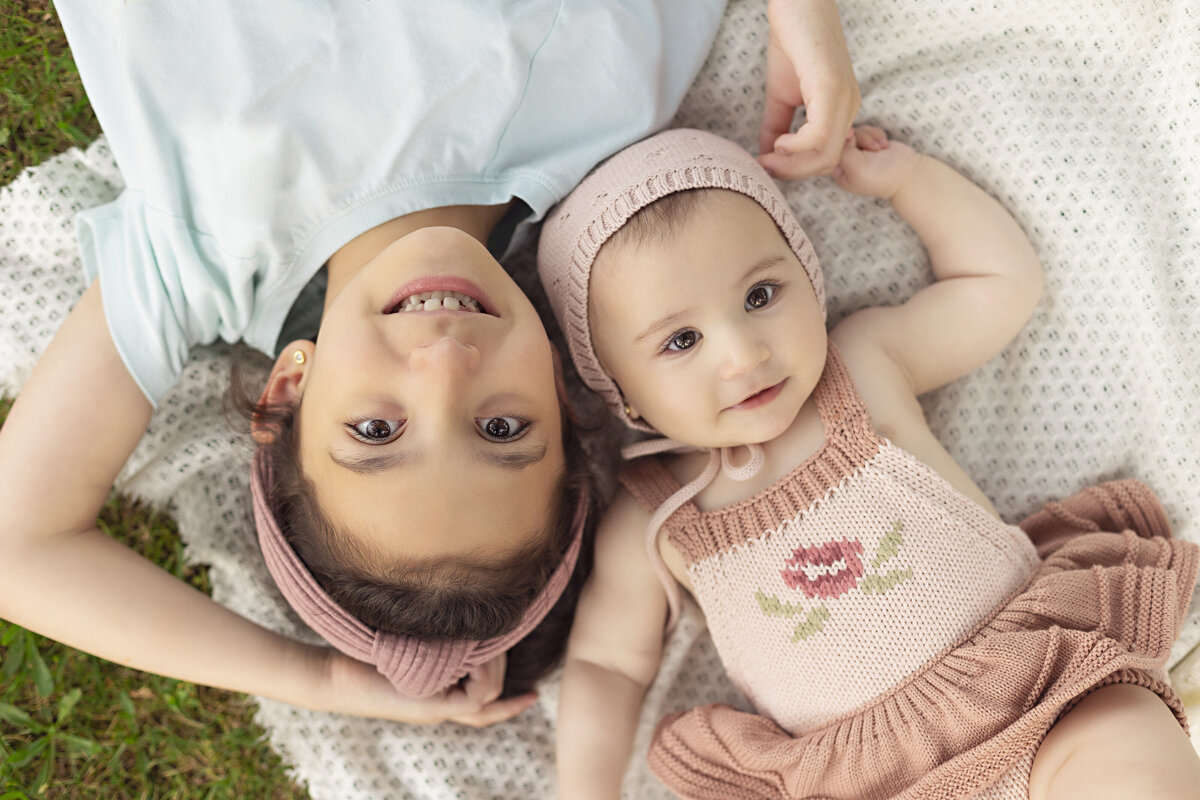 A young girl lays on a picnic blanket head to head with her infant baby sister in a rose embroidered dress onesie