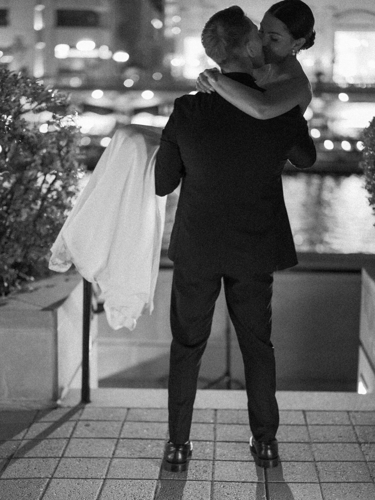 A cinematic black and white wedding photo of a bride and groom sharing a passionate kiss at night along the Chicago Riverwalk.