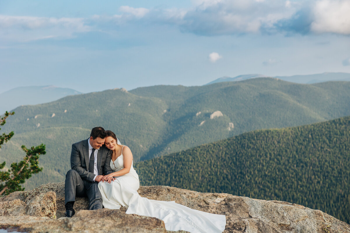 Couple is sitting on rocks together with a big overlook of the mountains. They are leaned in on each other. 