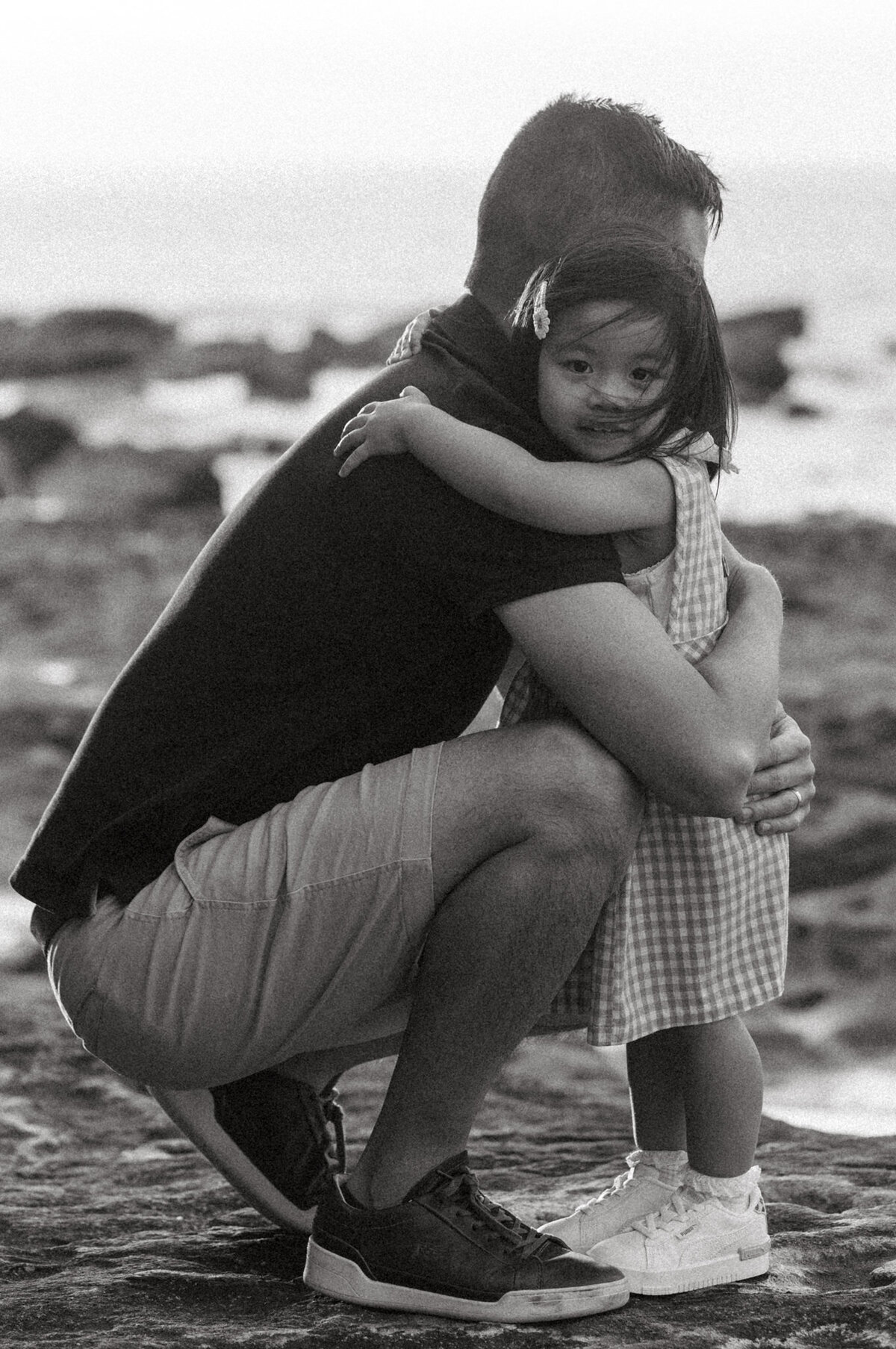 Black and white photo of a father crouching to hug his daughter during a beach family session, capturing a quiet and emotional connection.