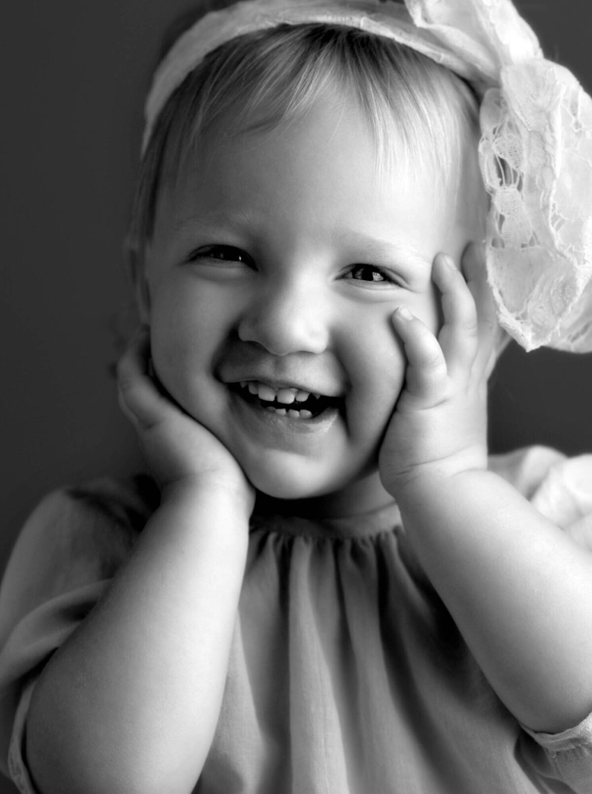 A joyful black-and-white portrait of a toddler with a lace headband, smiling widely and cupping their cheeks, conveying happiness and innocence.