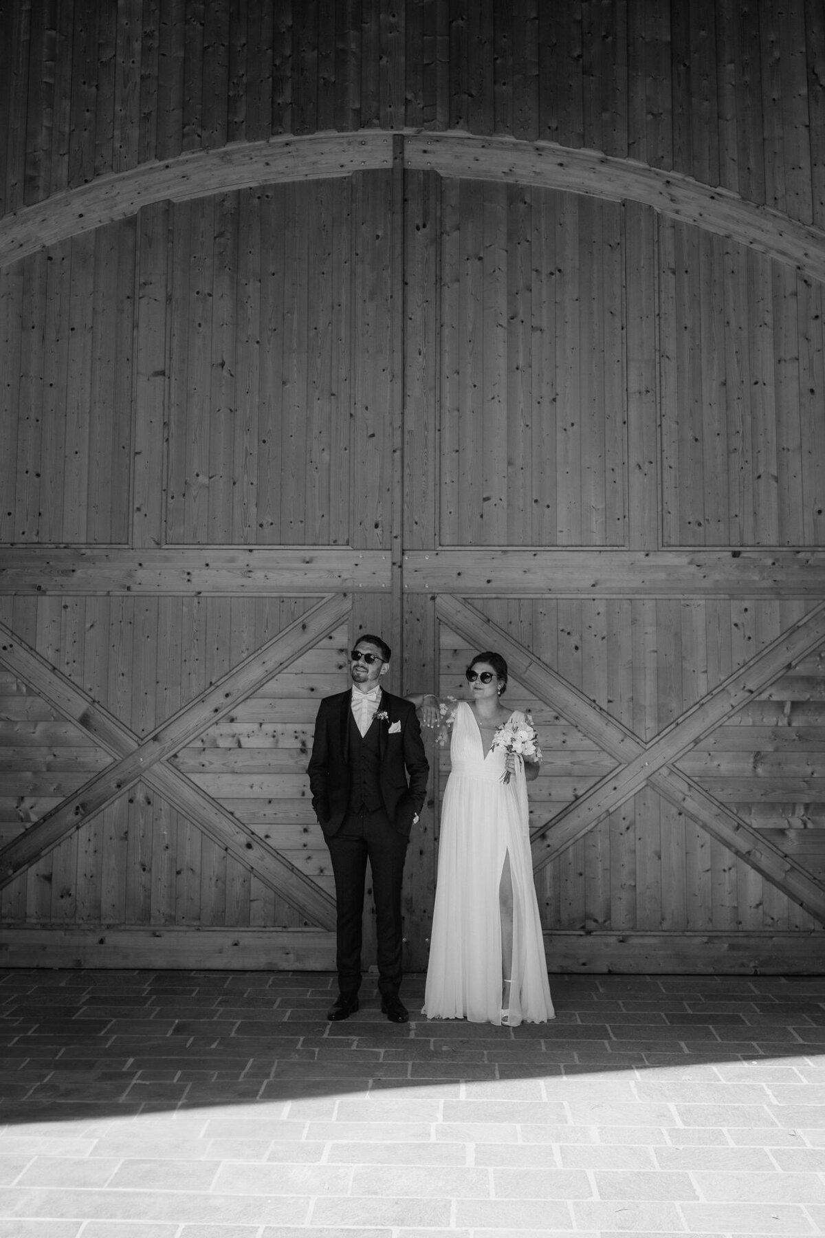 Das Hochzeitspaar steht vor einem großen Holztor in einer lässigen Pose nebeneinander. Beide tragen Sonnenbrillen.