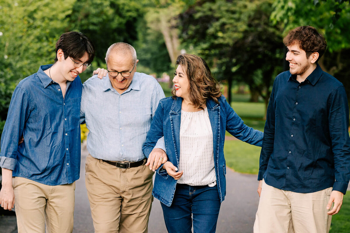 Two brothers in their early-twenties link arms with their older parents and are smiling and walking through the lush backdrop of a path in the Boston Common.