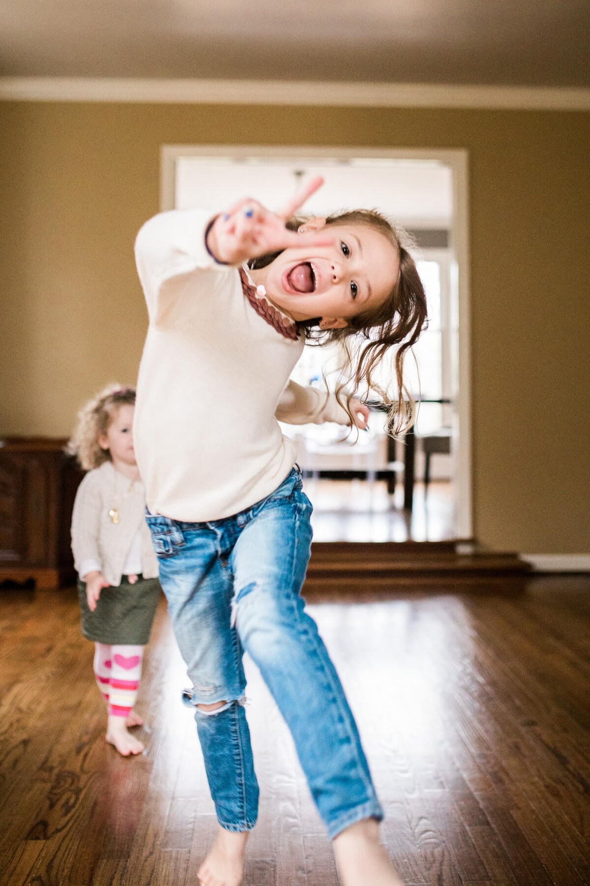 A young girl joyfully jumping in a living room, sticking out her tongue, with another child watching in the background during a DIY family photoshoot.