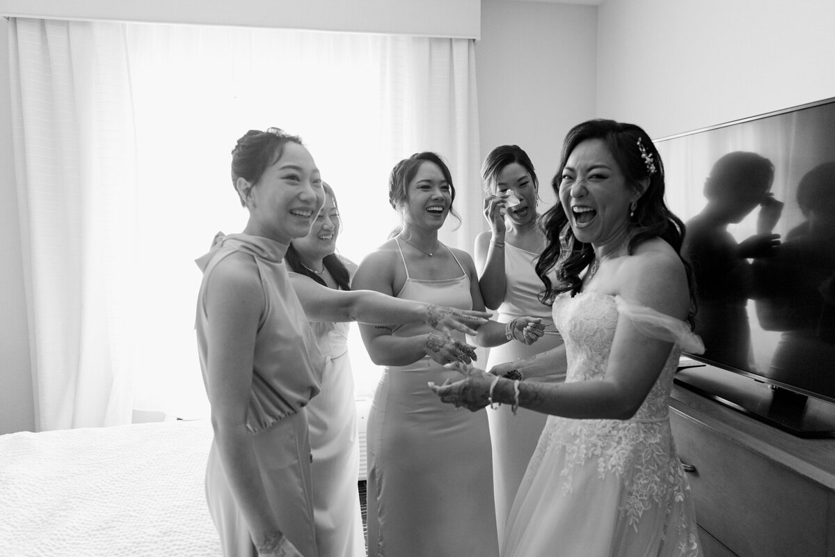 Black and white candid shot of a group of bridesmaids seeing the bride for the first time at a wedding in Dallas, Texas. Everyone is cheering, laughing, or wiping away tears. The bridesmaids are all in similar dresses and are happily reaching towards the bride. The bride is wearing an intricate white dress with a hairpiece and is laughing enthusiastically.