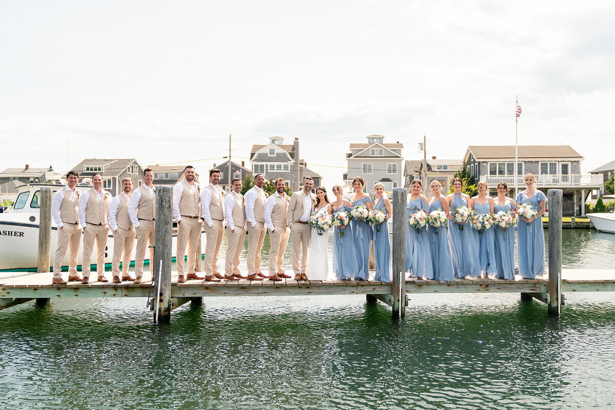 A wedding party lined up on a dock, groomsmen in beige suits and bridesmaids in blue dresses, with waterfront houses in the backdrop.