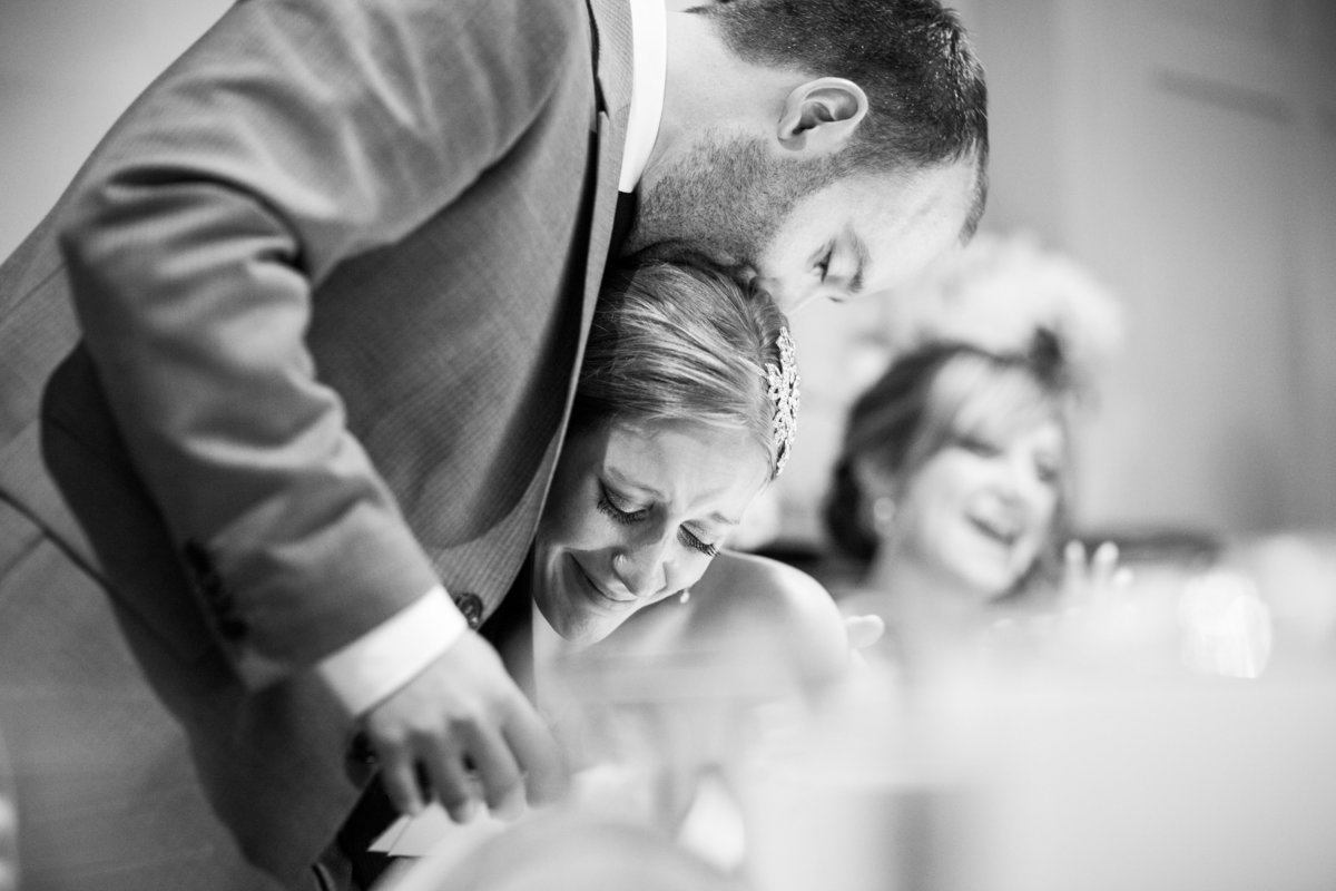 black and white reportage photo of the groom kissing the brides head in the wedding speeches